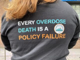 The back of a sweater with slogan reading "every overdose death is a policy failure"